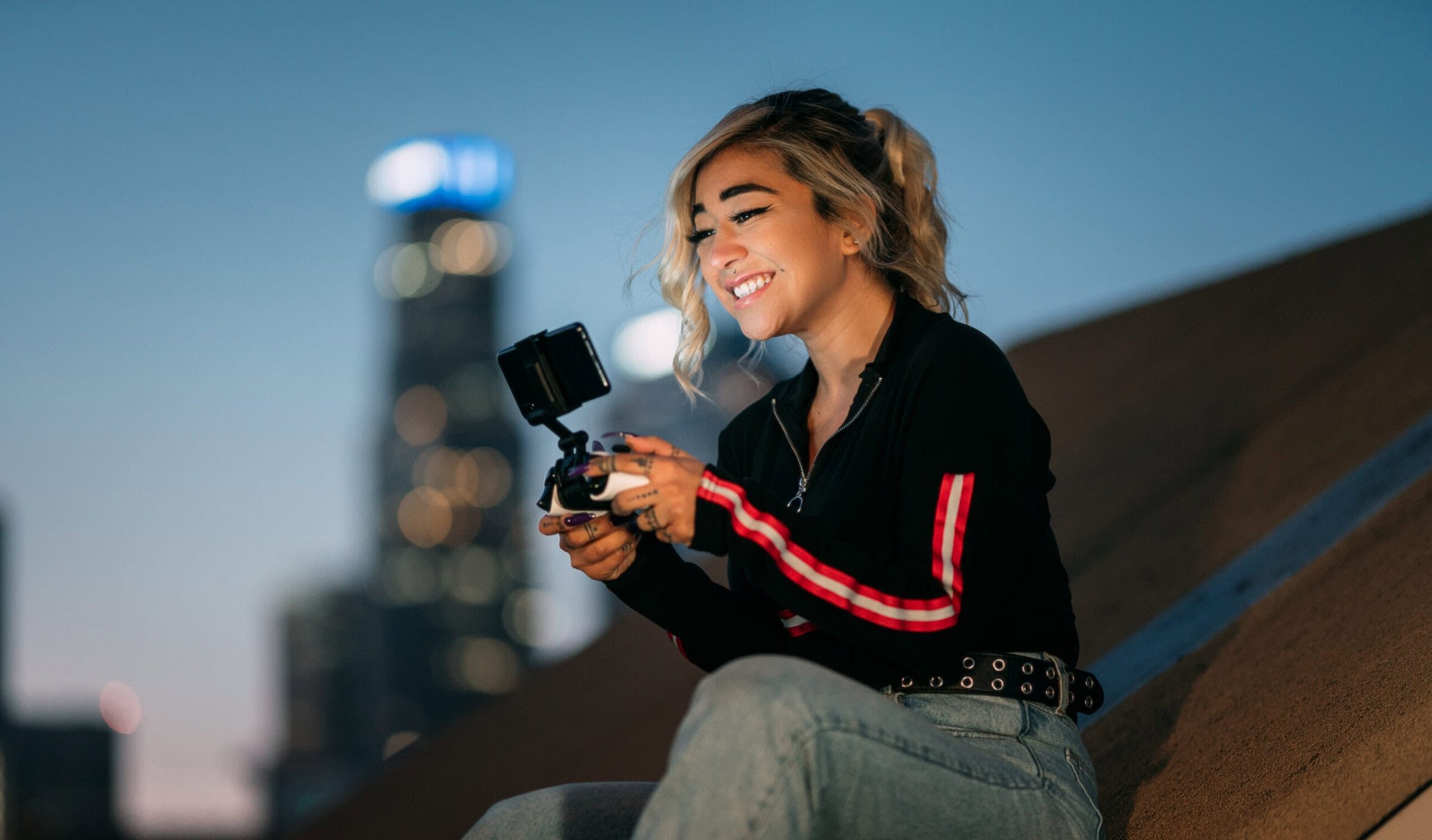 Young woman with blonde hair smiles while holding a camera on a rooftop at dusk. She's wearing a black jacket and jeans. Blurred city lights in background.