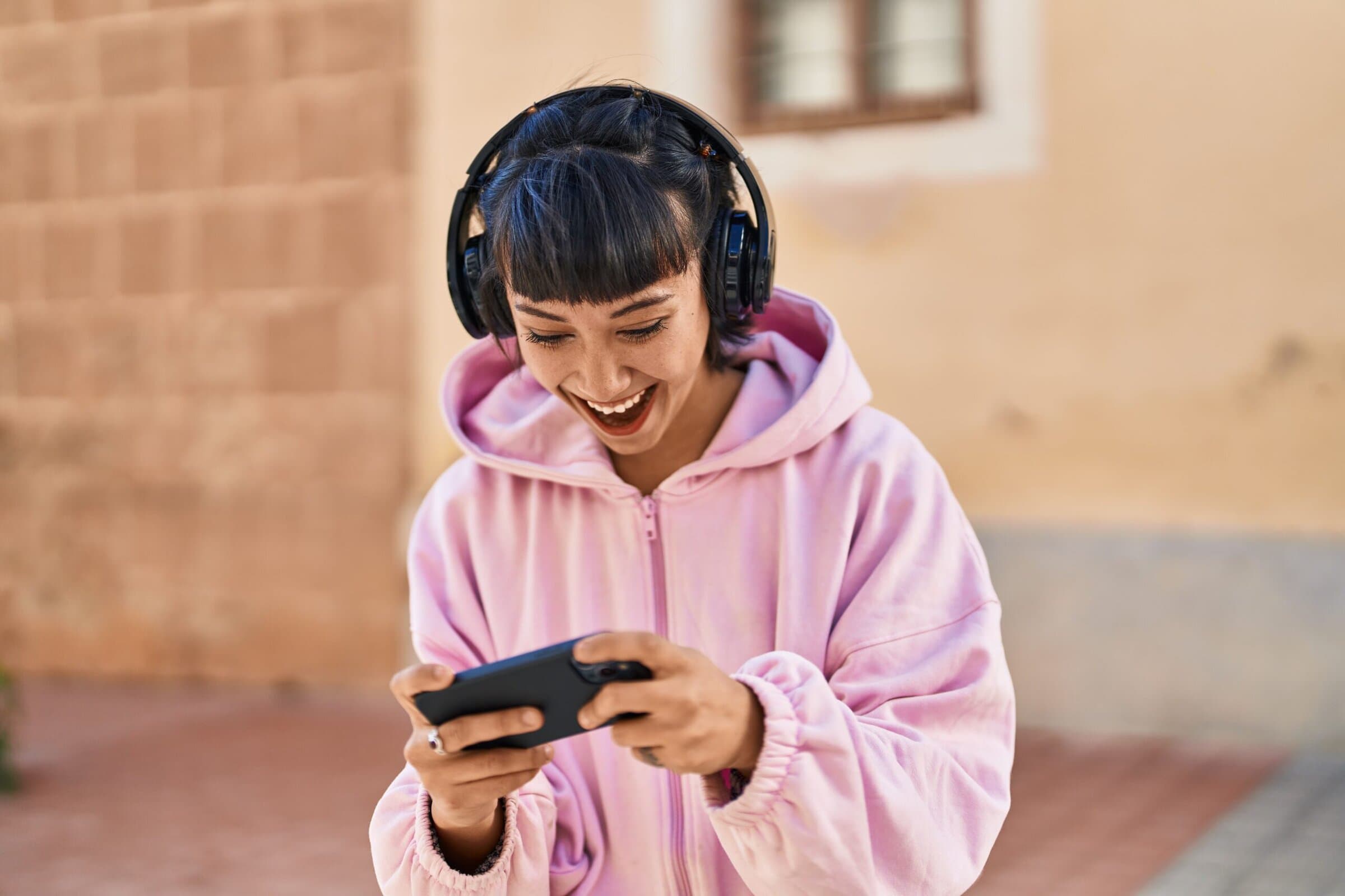 A person in a pink hoodie, wearing headphones, is smiling while looking at a smartphone. The background shows a textured wall, conveying a joyful mood.
