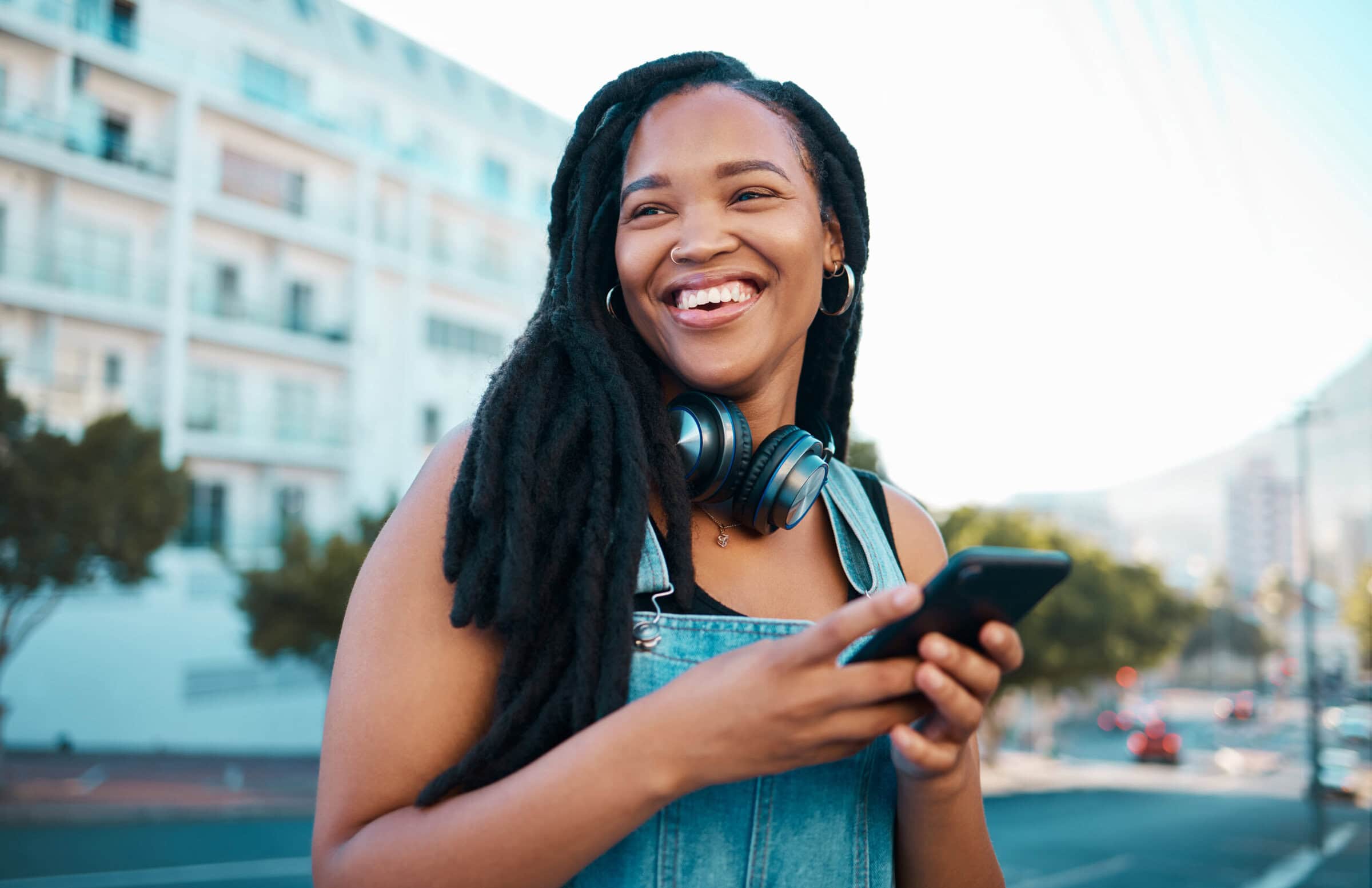 Smiling woman with headphones around her neck holds a smartphone, standing outdoors in front of a blurred building and trees, exuding happiness.