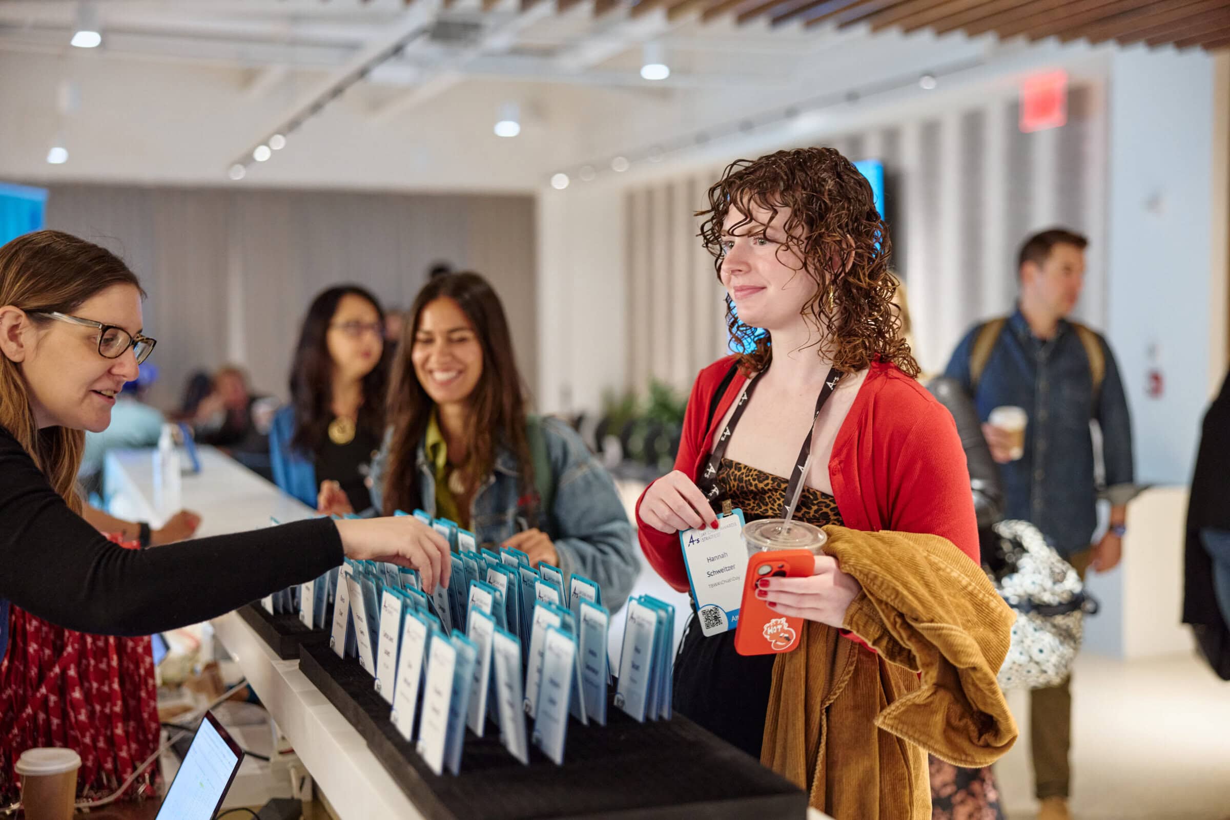 A woman in a red top smiles while picking up a name badge from a table of name tags. Two people stand nearby, and a casual atmosphere is conveyed.