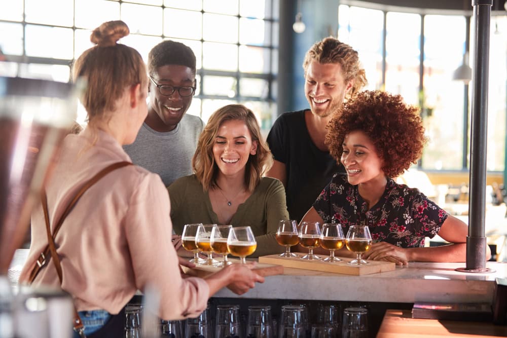 A group of five friends at a bar, smiling and eagerly receiving a tray of beer samples from a bartender. The atmosphere is bright and cheerful.