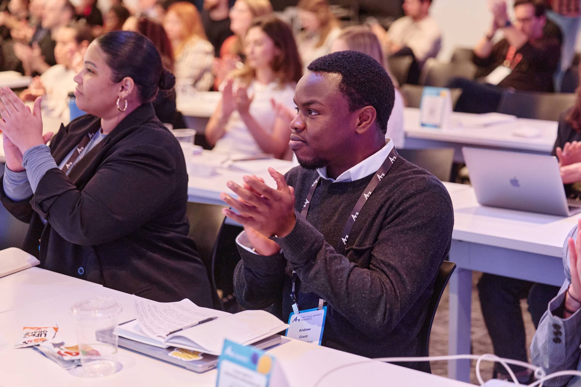 People seated at a conference, attentively clapping. The setting is a well-lit room with notebooks and laptops on tables, conveying focus and engagement.