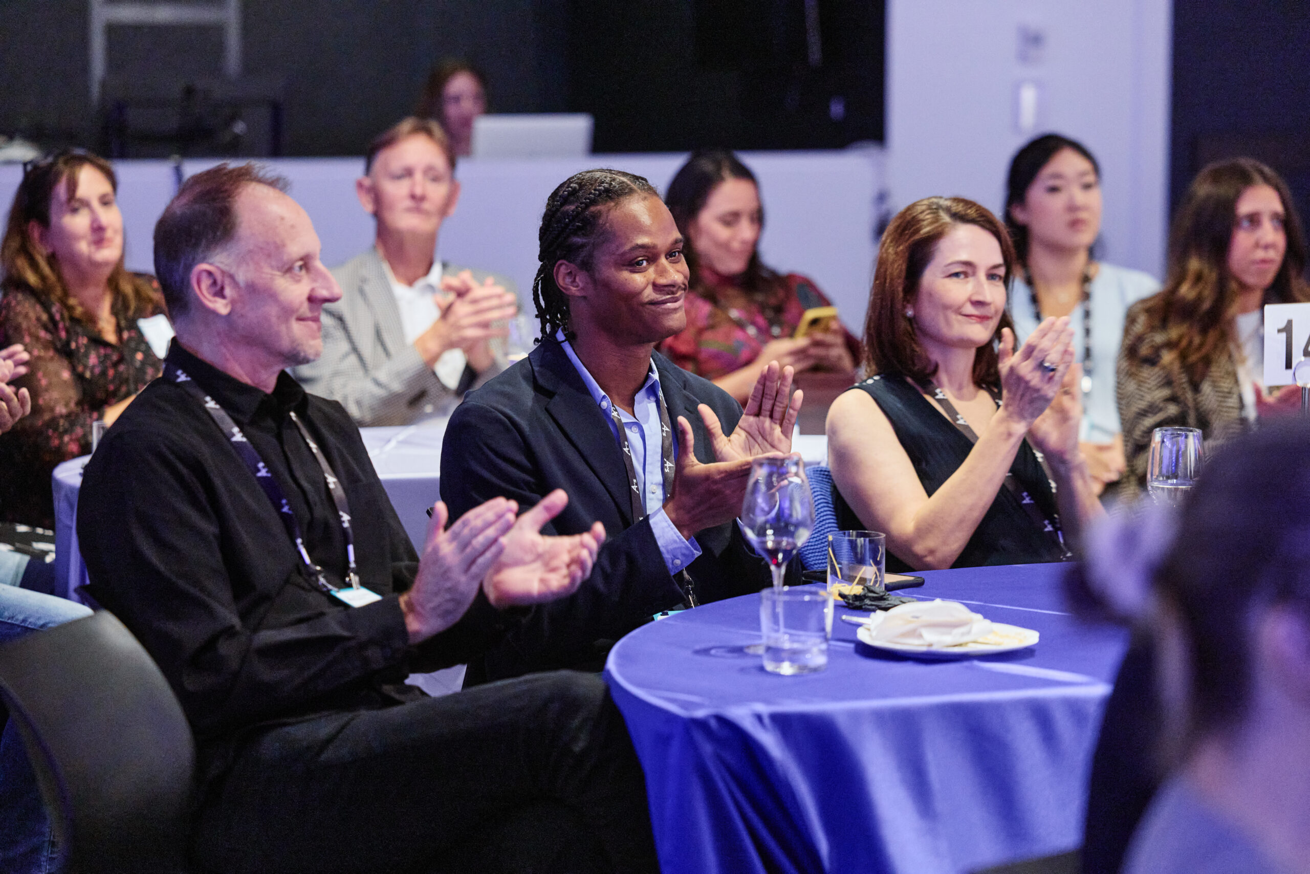 A group of people sitting at a table with blue tablecloths, attentively clapping during an event. The mood is engaged and positive.