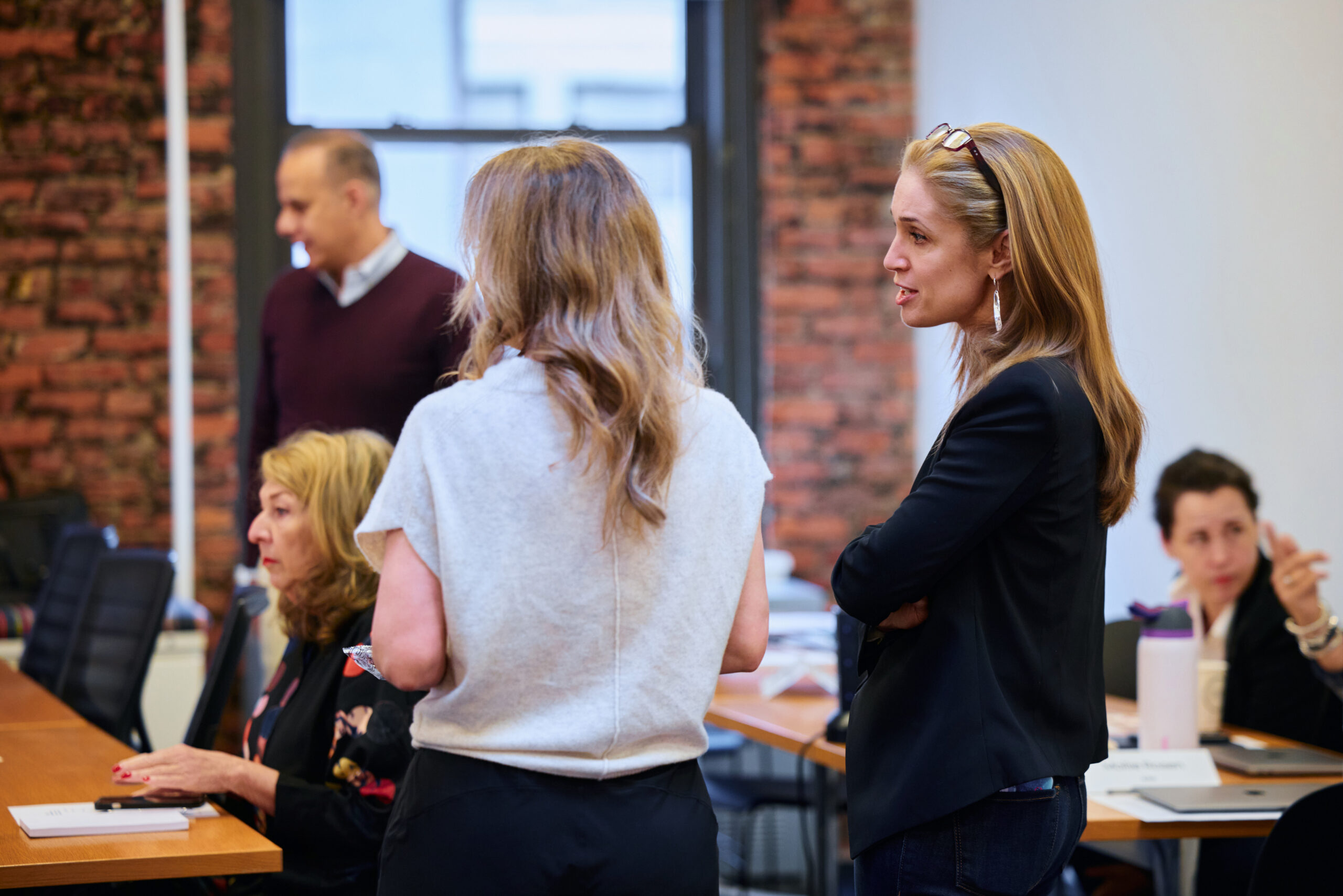 A group of people in a meeting room engage in conversation around a table. The mood is collaborative, with a brick wall and window in the background.