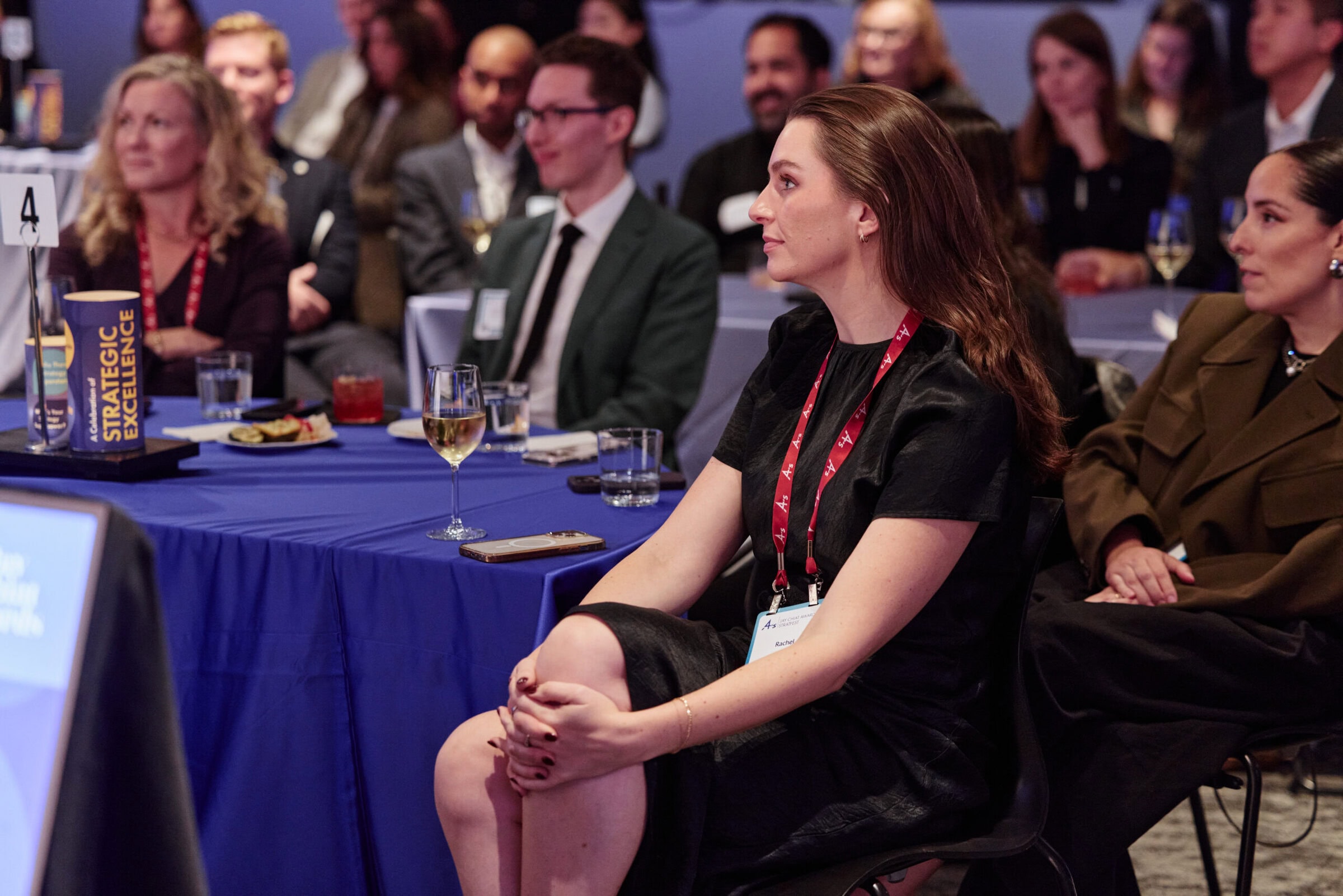 A woman in a black dress sits attentively at a conference table with a blue tablecloth. She is surrounded by engaged attendees, creating a focused atmosphere.