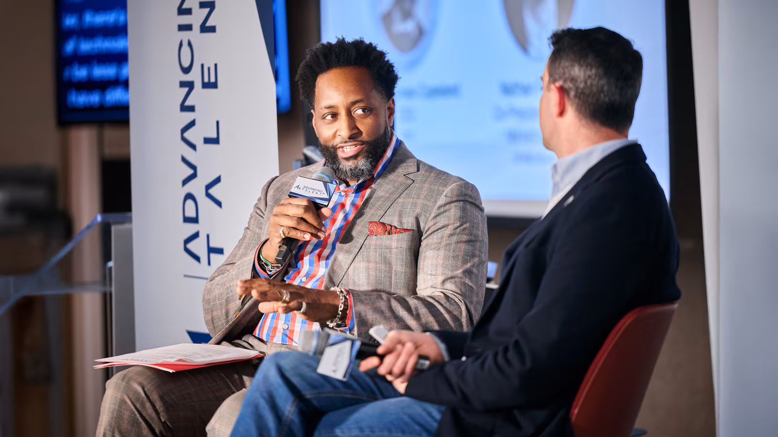Two men sitting in chairs talking on a stage.