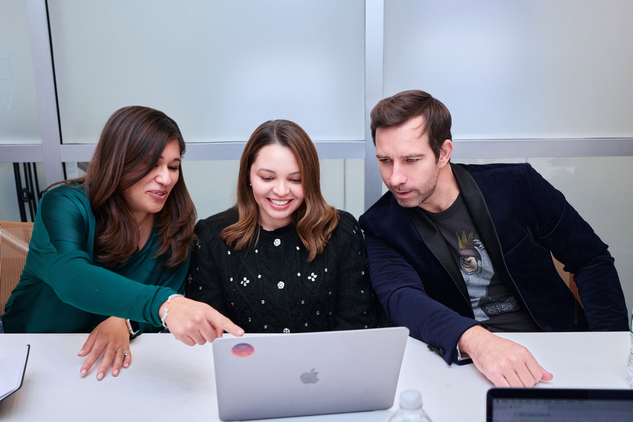 Three people sit at a table, smiling and engaged, looking at a laptop. The woman on the left points at the screen, suggesting teamwork and collaboration.