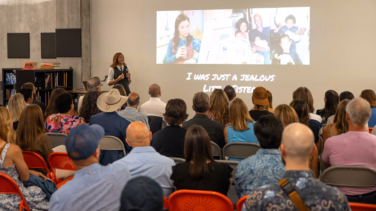 A woman presents to an audience in a modern room, showing personal photos and text on a large screen. The slide reads "I was just a jealous little sister." The audience is seated attentively, conveying interest.