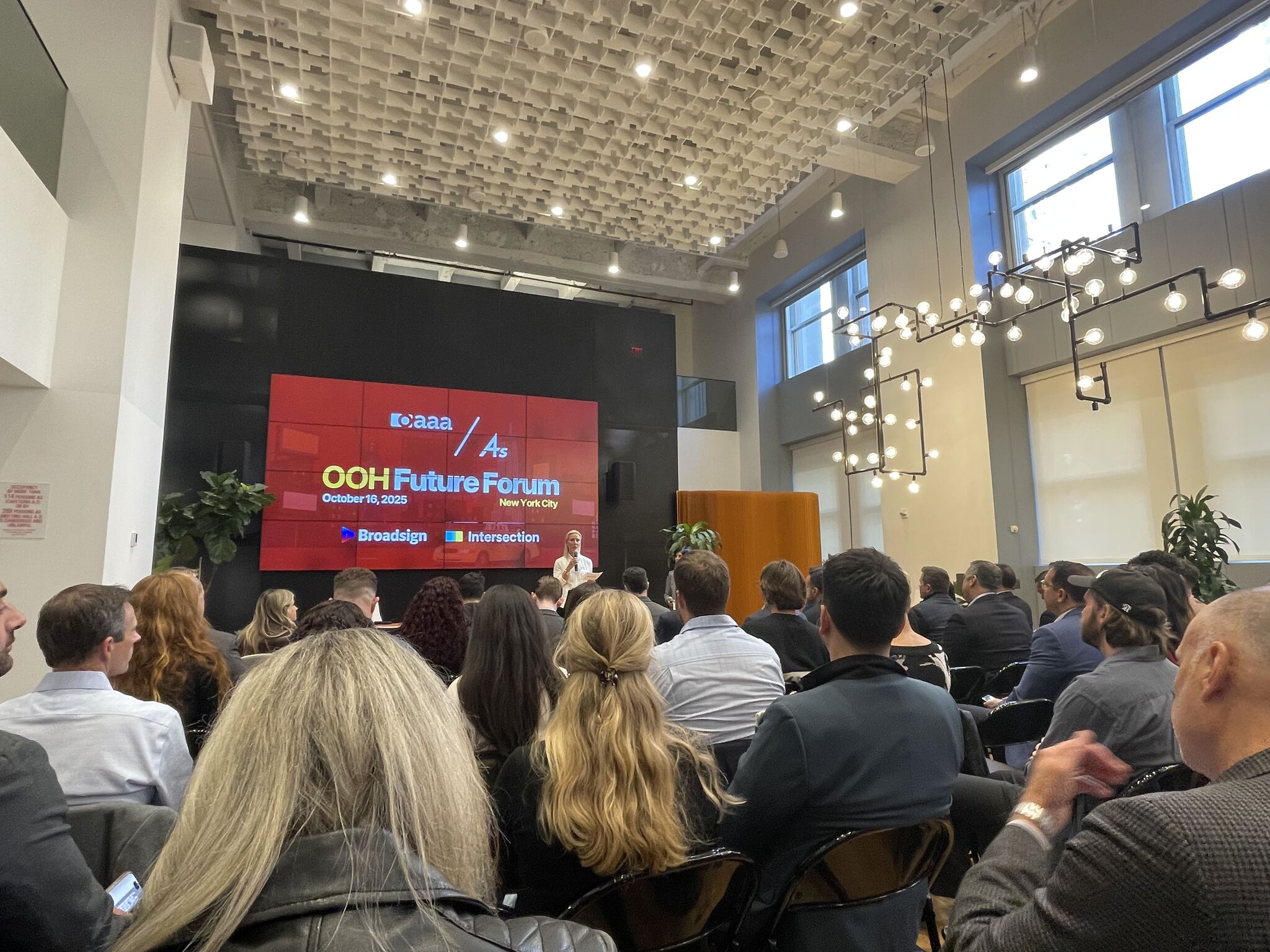 Audience watching a speaker at the "OOH Future Forum" in New York City, with a large red screen and modern, bright room ambiance.