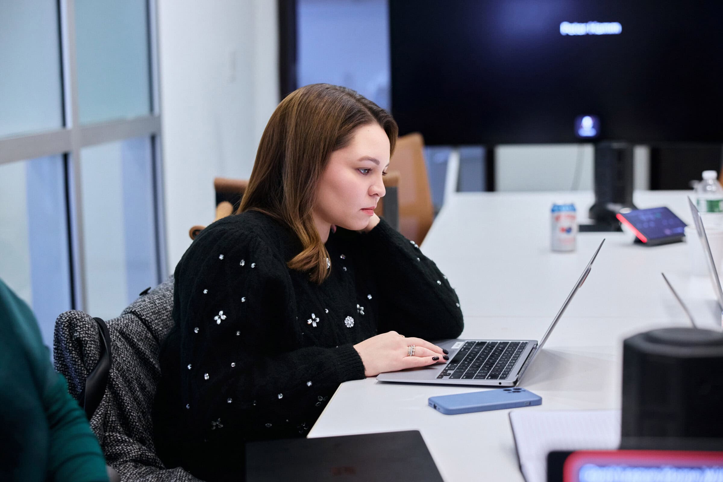 A woman in a black sweater sits at a conference table, focused on her laptop. The room is bright and modern, creating a professional atmosphere.