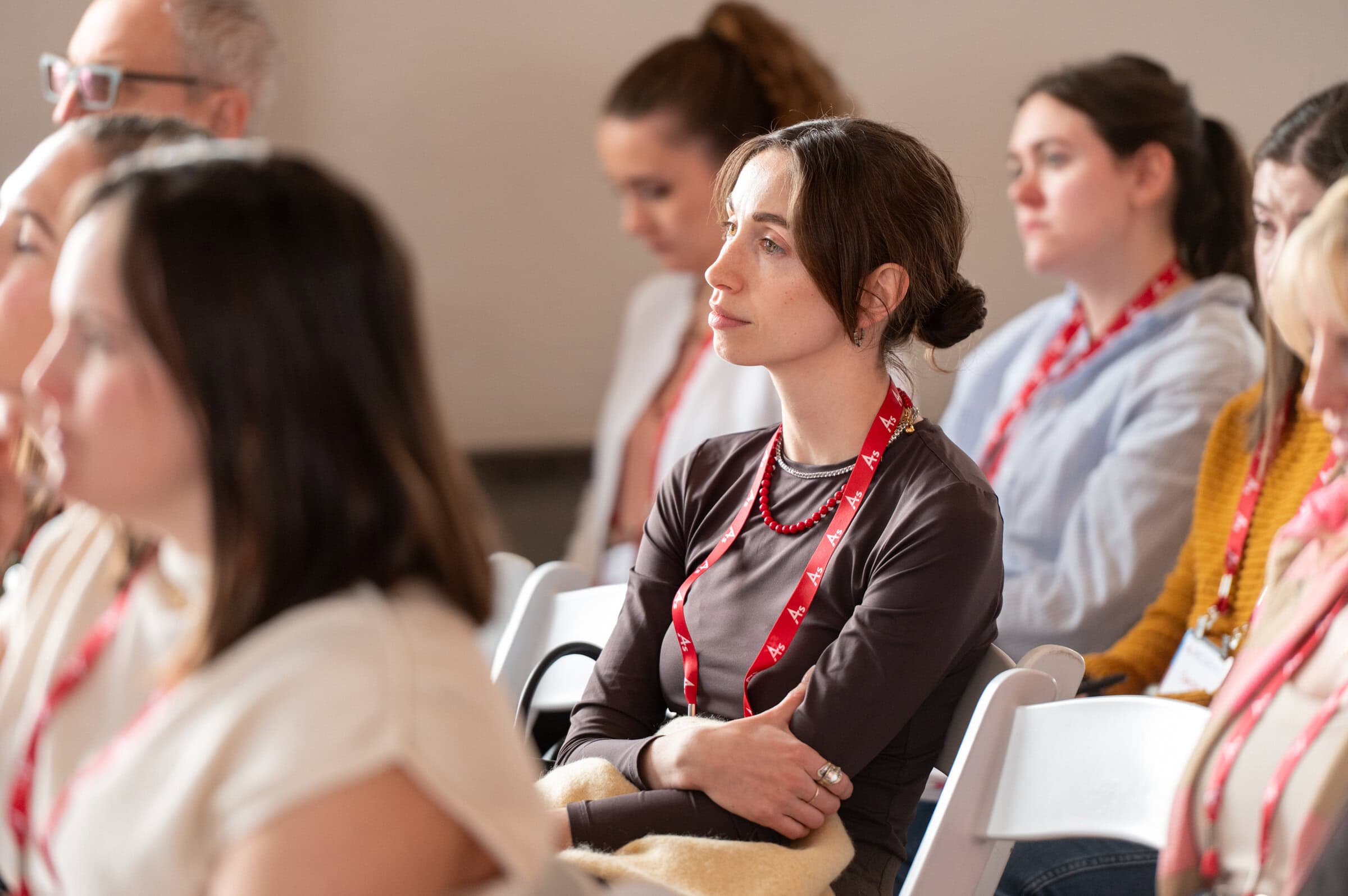 A group of attentive people sitting on white chairs, wearing lanyards, focuses on a speaker outside the frame. The mood is thoughtful and engaged.