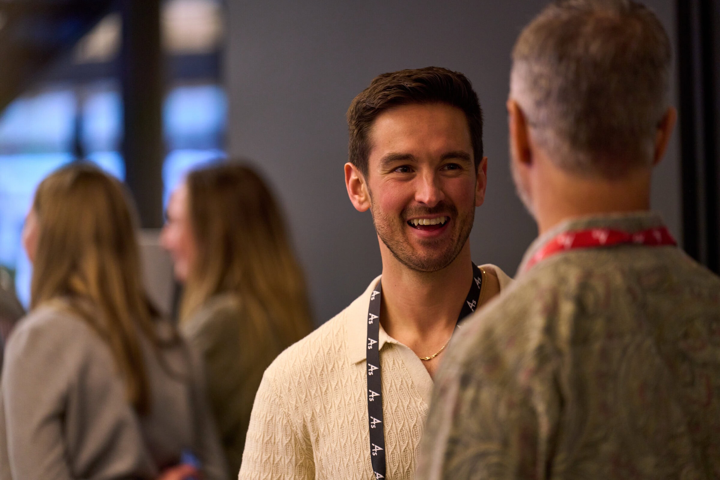 A man in a white sweater with a lanyard smiles while talking to another man in a patterned shirt. Blurred people are in the background, creating a warm, friendly atmosphere.