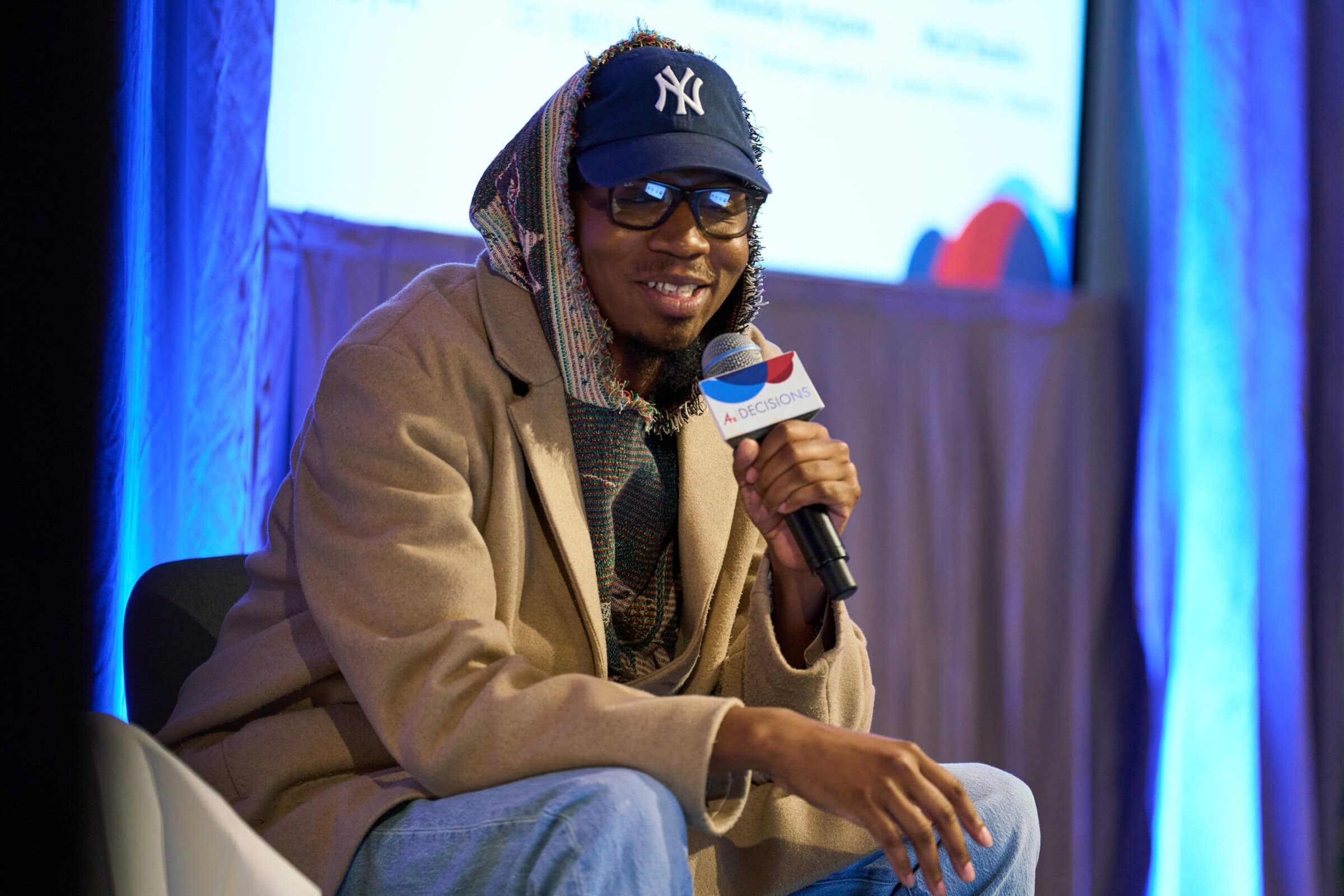 A person wearing a New York Yankees cap, glasses, and a beige coat sits on stage holding a microphone, smiling. The background is softly lit in blue.