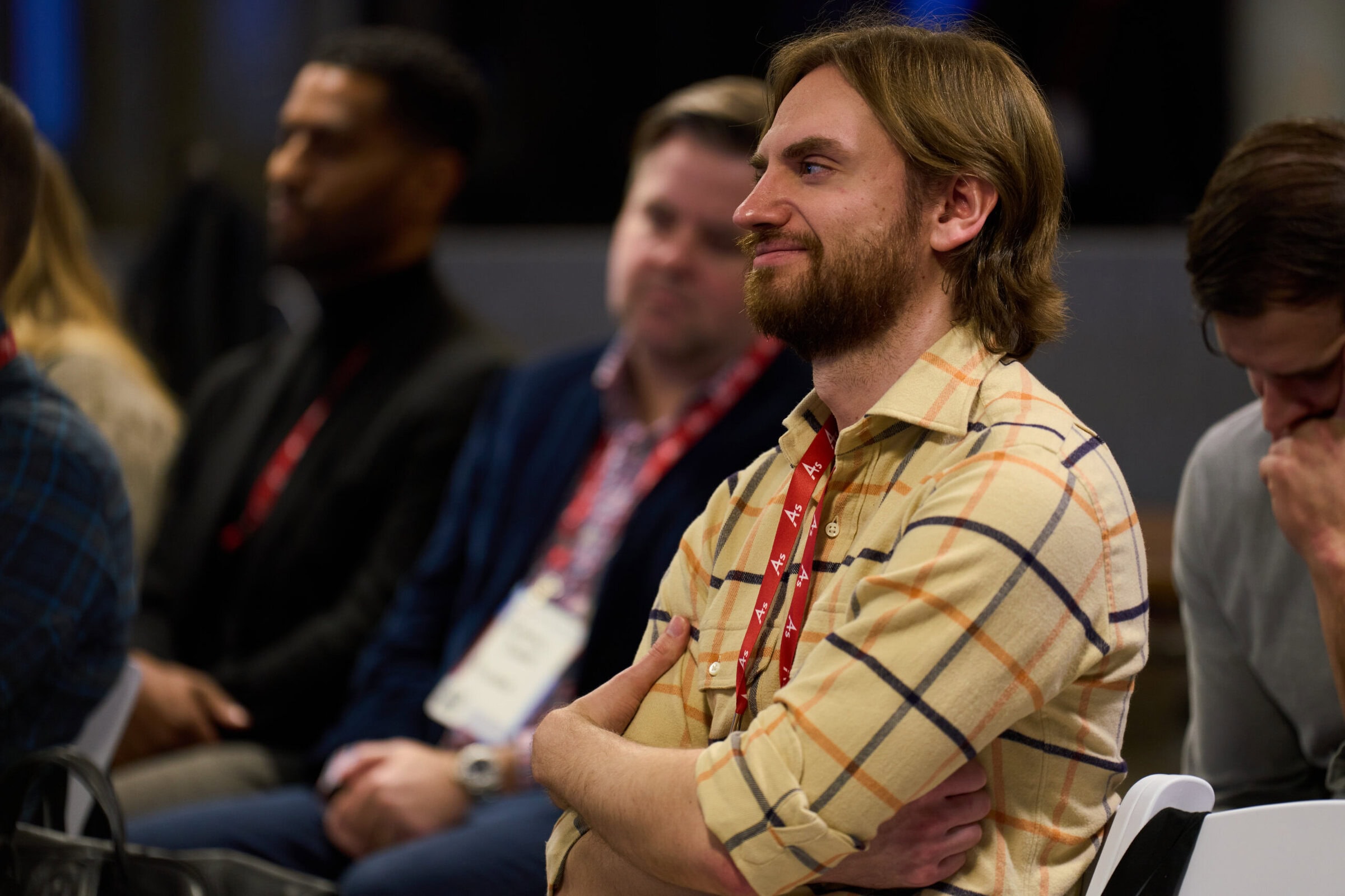 A man with a beard, wearing a plaid shirt, smiles and crosses his arms while seated at an event. People in the background appear attentive and engaged.