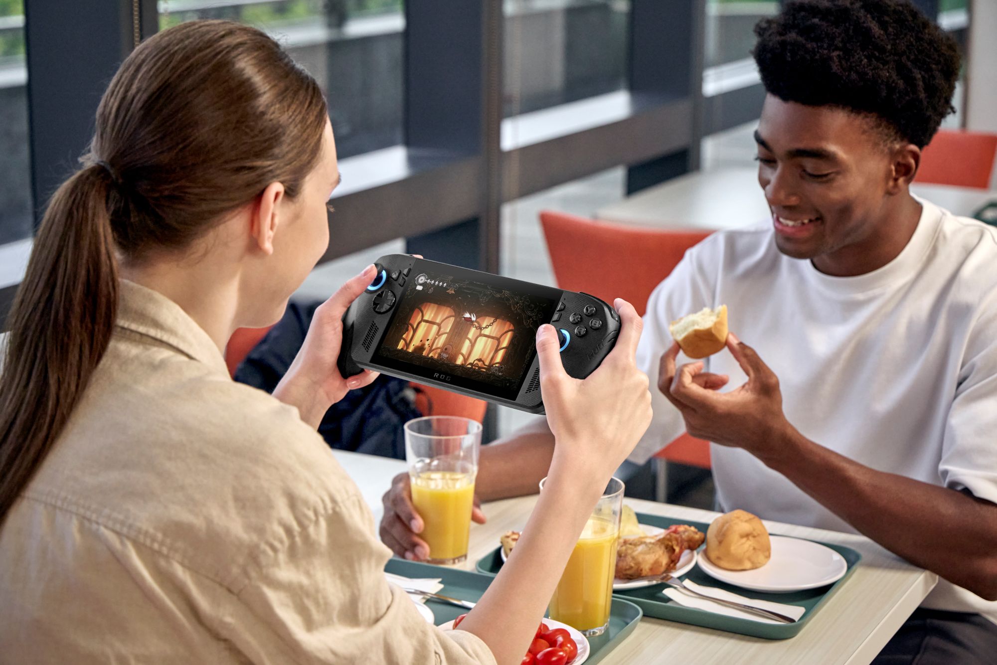 A woman plays on a handheld gaming device, showing a game screen, while a man smiles and holds a sandwich. Bright, casual dining setting with trays of food.