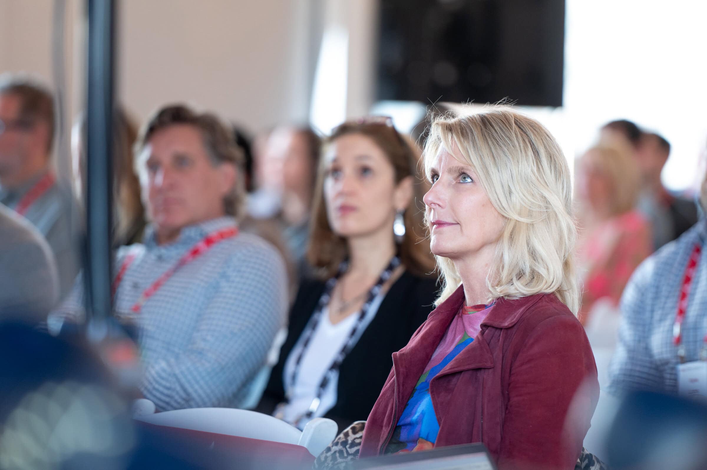 A focused, attentive woman with blonde hair and a red jacket listens in a seminar room. Blurred attendees, wearing lanyards, fill the background.