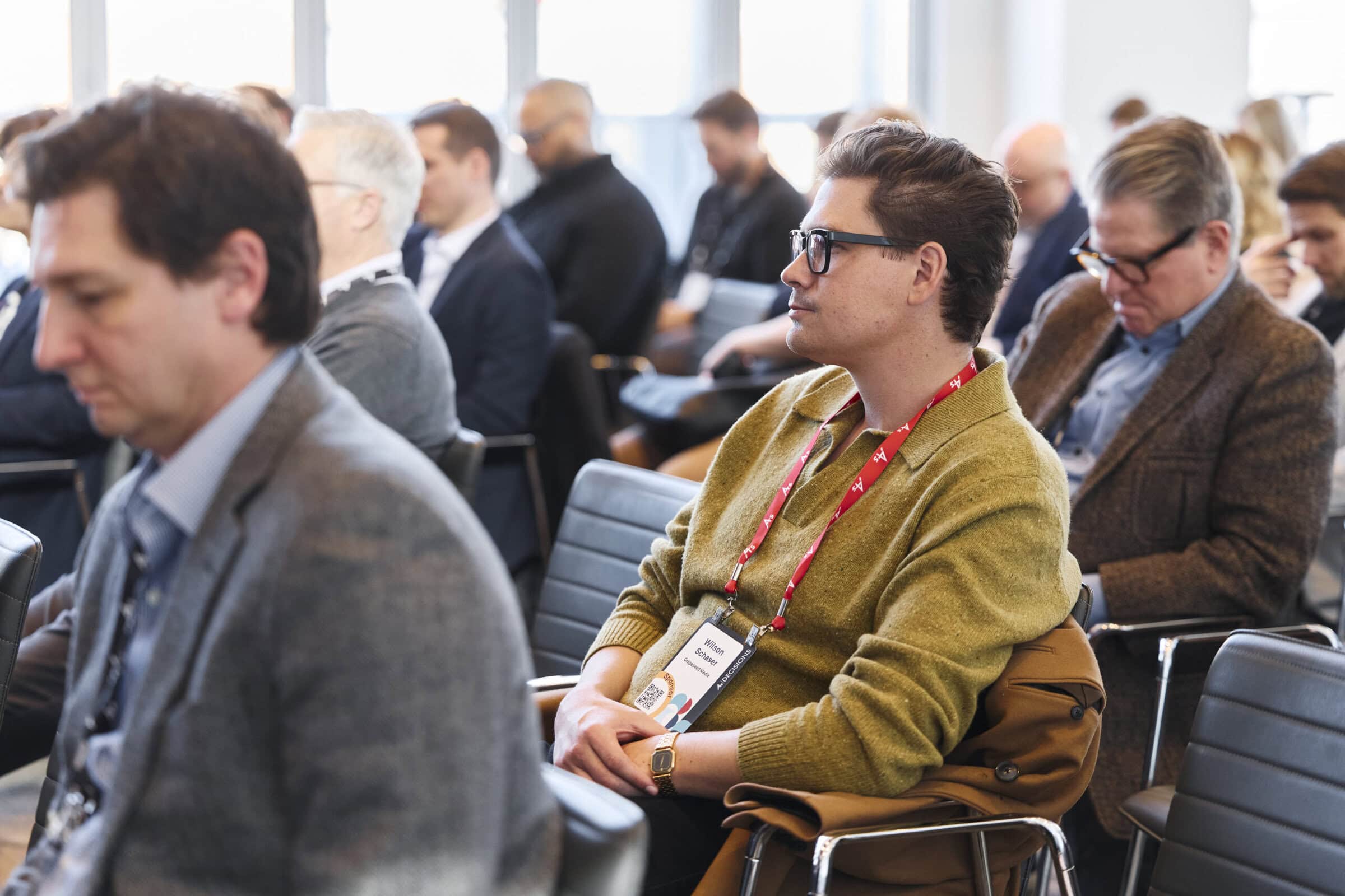 A conference room filled with attentive attendees sitting in rows. The focus is on a man wearing glasses and a green sweater. The atmosphere is focused and professional.