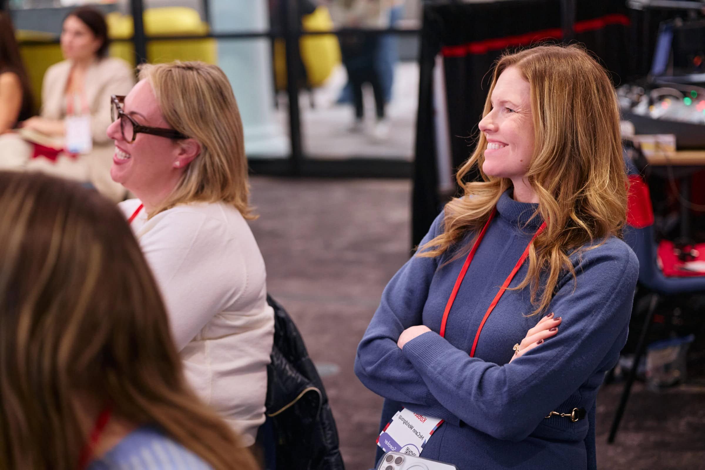 Two women with conference badges smile and watch attentively. They appear engaged and happy in a bright, professional setting.