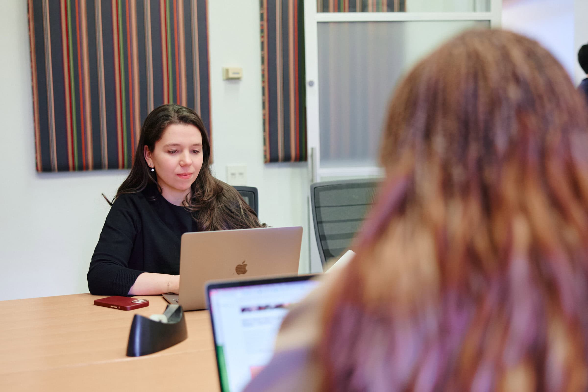 A woman in a conference room works on a laptop, focused and engaged. Another person with blurred red hair is in the foreground. The setting is professional.