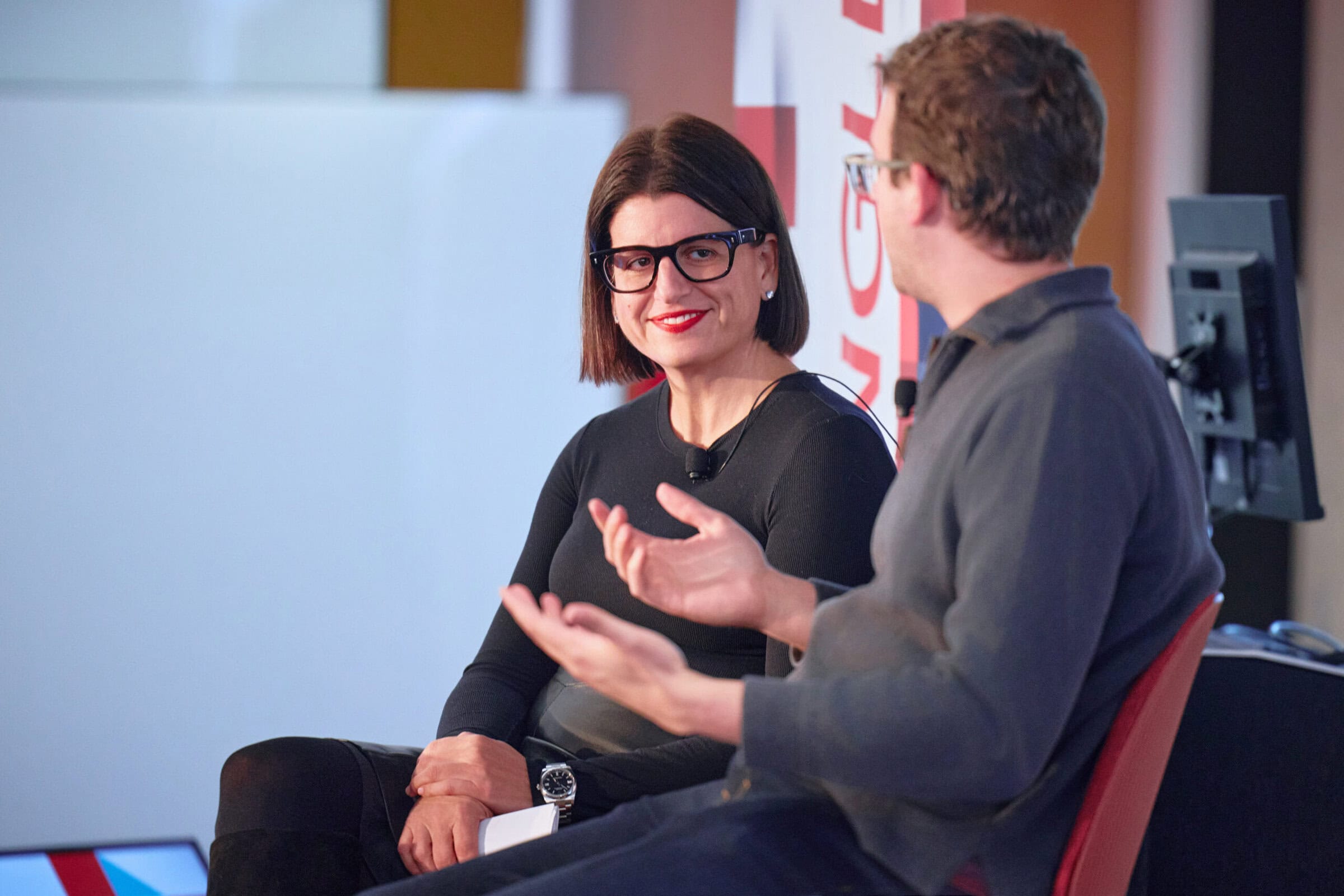 Two people seated and engaged in conversation during a conference. One is smiling, wearing glasses, and a black sweater; the other gestures while speaking.