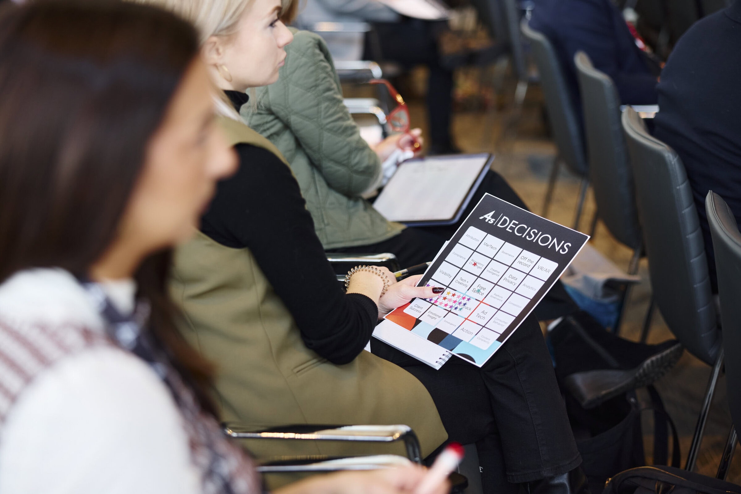 A group of people sit in rows of chairs during a conference. A woman in focus holds a "Decisions" brochure. The setting appears professional and focused.
