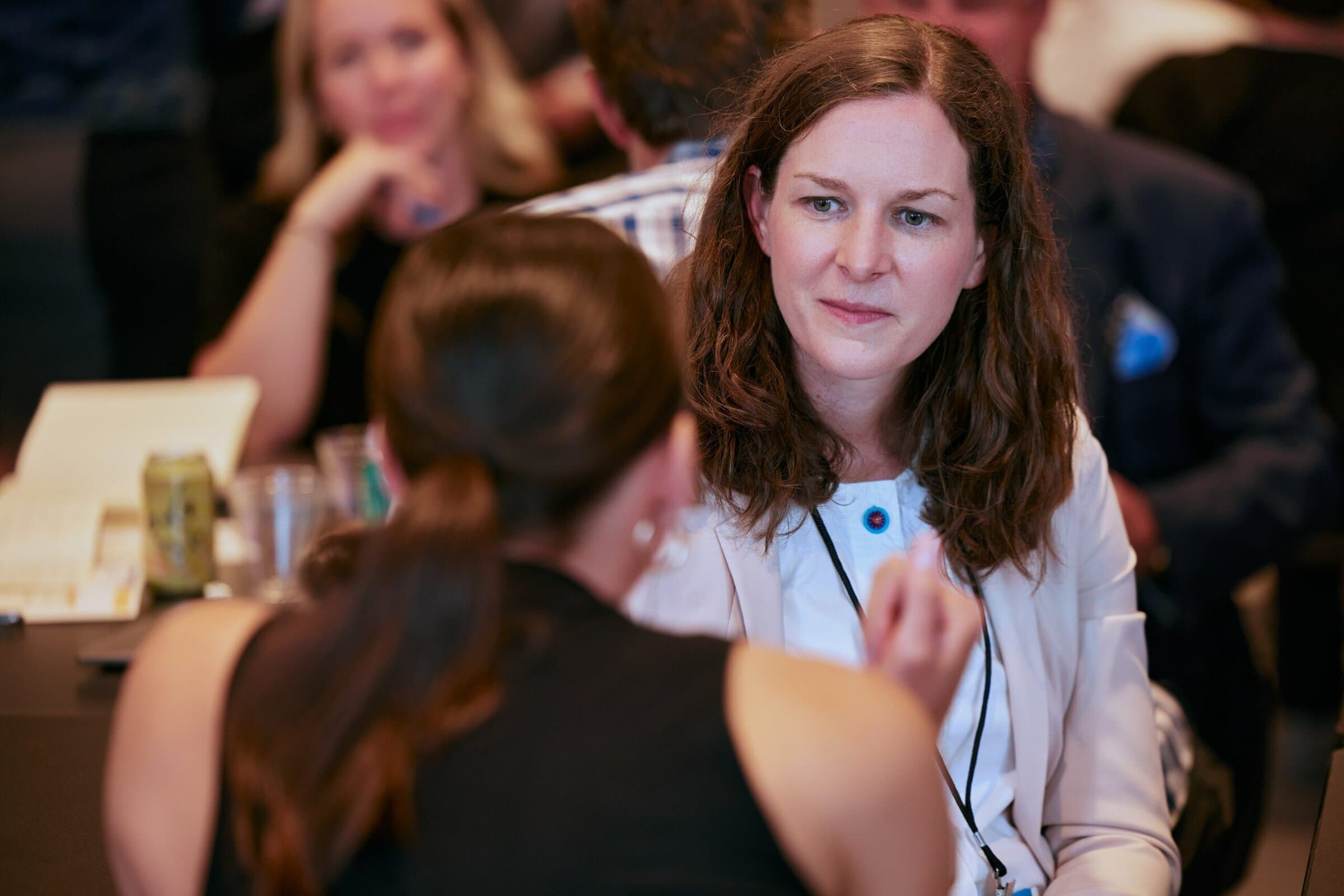 A woman with long hair listens intently during a conversation at a conference. She wears a white blazer, conveying a professional tone.