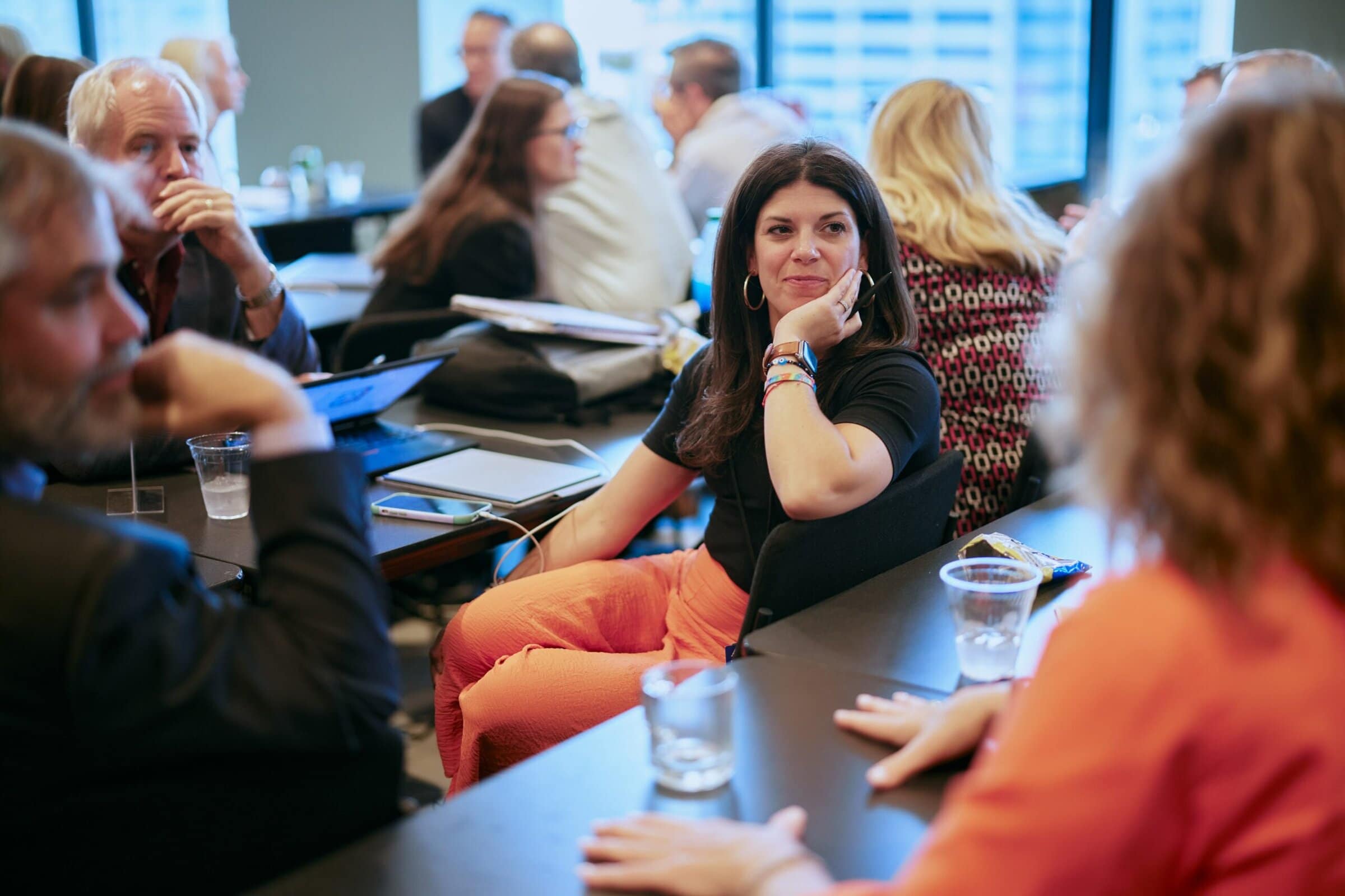 A woman in orange pants and a black shirt engages in a lively group discussion at a conference table. The atmosphere is collaborative and focused.