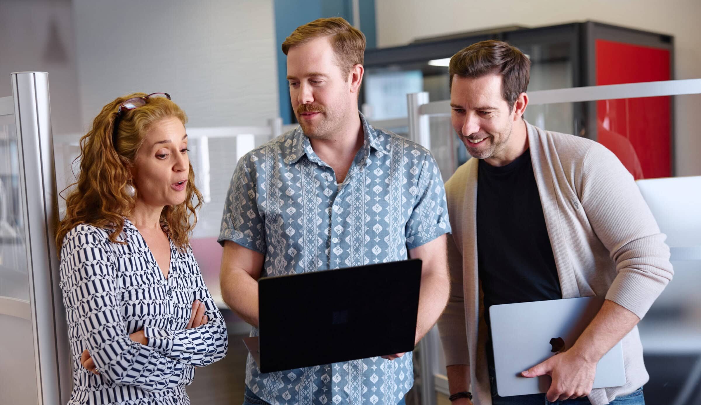 Three people in a casual office setting, engaged in discussion. A woman with curly hair is talking to two men holding laptops, all appear focused and collaborative.
