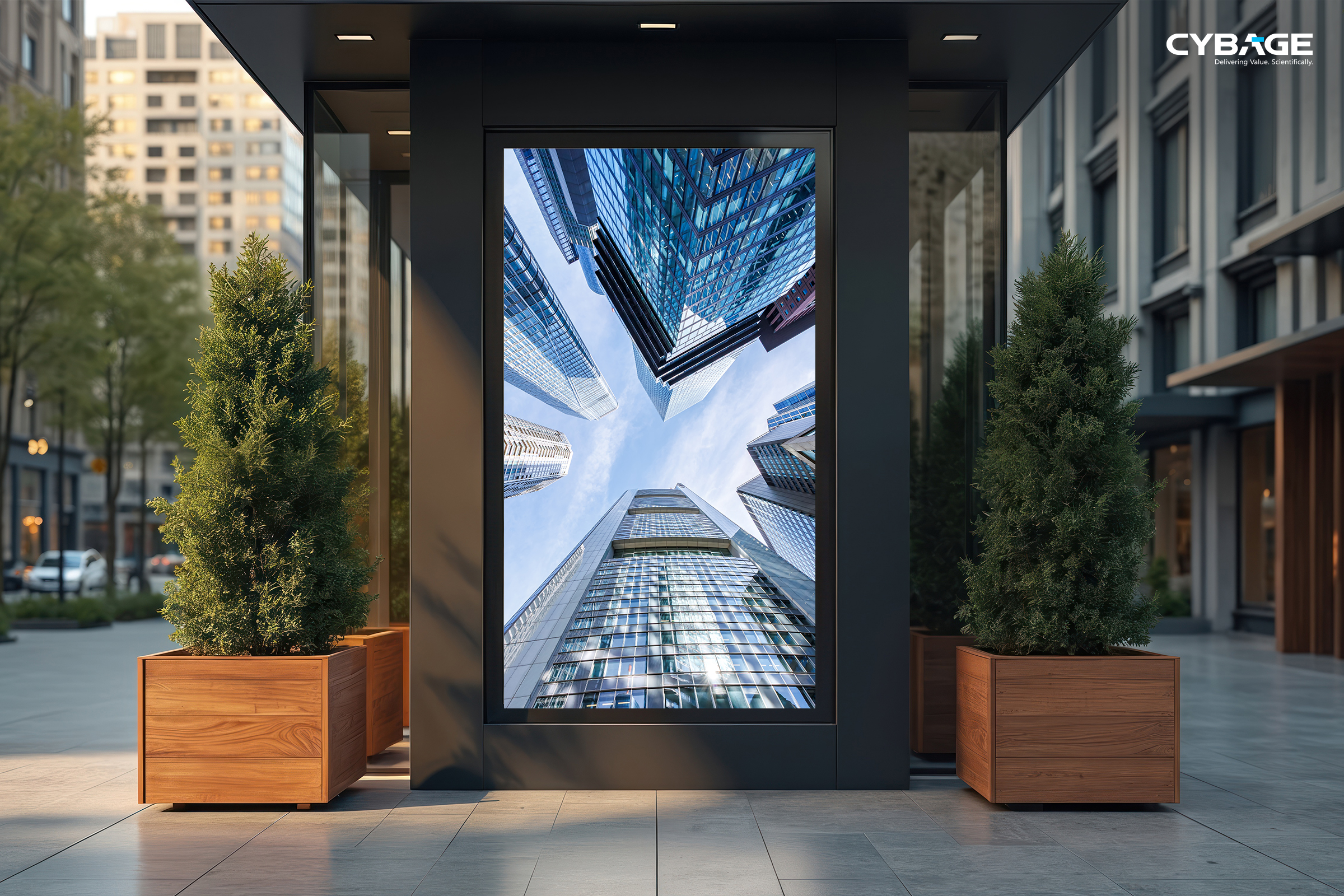 Vertical digital display in a modern urban setting shows skyscrapers from below against a blue sky. Framed by wooden planters with green shrubs.