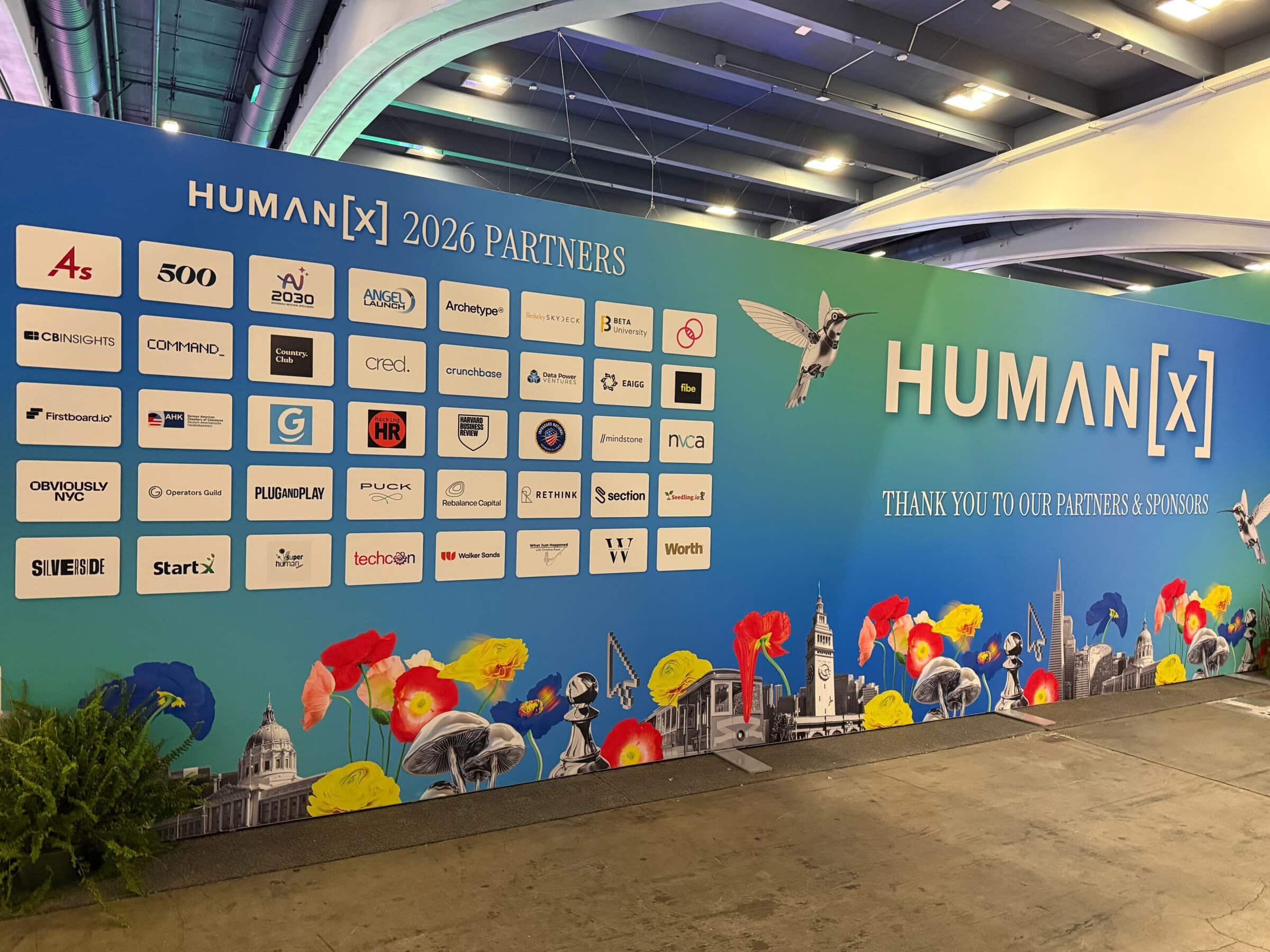 A vibrant display showcases "HUMAN[X] 2026 Partners" logos on a blue wall with colorful flowers. The scene has a dynamic and welcoming atmosphere.