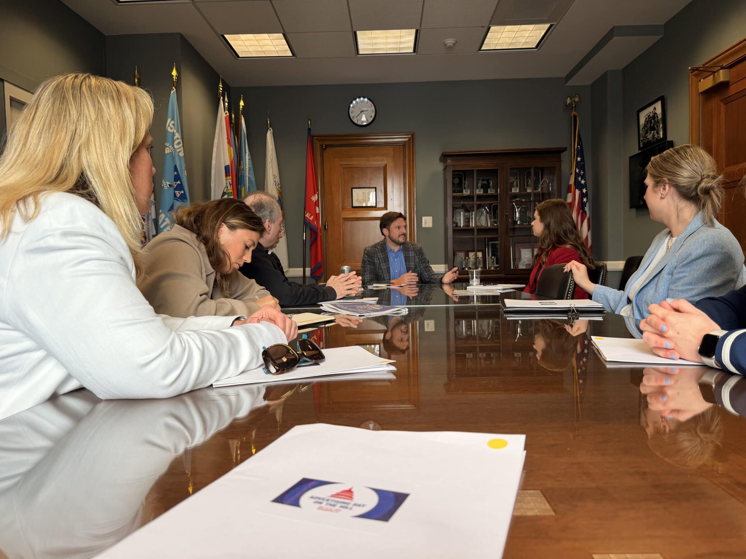 A group of six people in a conference room engage in a discussion around a polished wooden table with papers, flags, and a clock on the wall.