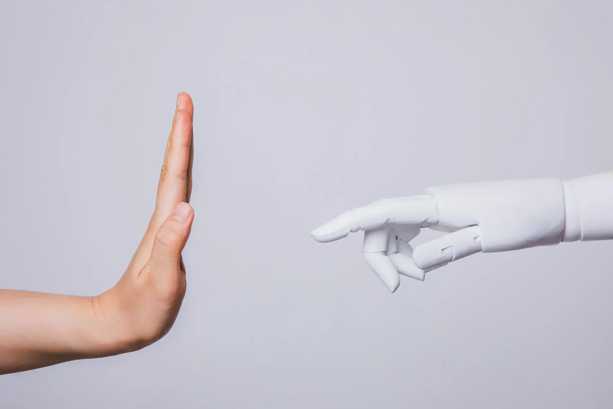 A human hand raised in a stop gesture faces a robotic hand reaching out. The neutral background emphasizes the contrast between human and technology.
